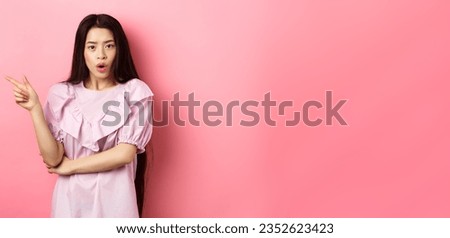 Shocked asian teen girl in dress pointing finger left, gasping wondered and look curious at camera, asking question about promo, standing on pink background.
