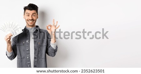 Image of excited man holding dollar bills and show okay sign with happy face, making money, standing on white background.