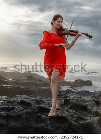 Attractive woman with violin on the beach. Music and art concept. Slim girl wearing short red dress and playing violin in nature. Sunset time. Cloudy sky. Bali, Indonesia