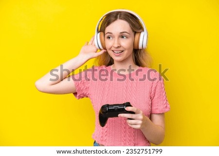 Blonde English young girl playing with a video game controller isolated on yellow background listening to something by putting hand on the ear
