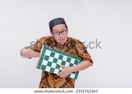 An awkward indonesian man inviting someone to play a game of chess. Holding a full-sized chessboard. Wearing a batik shirt and songkok skull cap. Isolated on a white background.