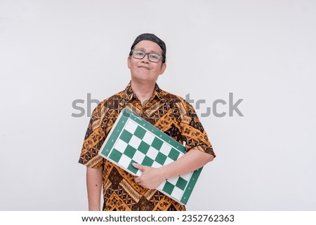 An indonesian man holding a full-sized chessboard. An amateur or professional chess player at a tournament. Wearing a batik shirt and songkok skull cap. Isolated on a white background.