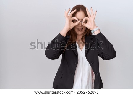Beautiful brunette woman wearing business jacket and glasses doing ok gesture like binoculars sticking tongue out, eyes looking through fingers. crazy expression. 
