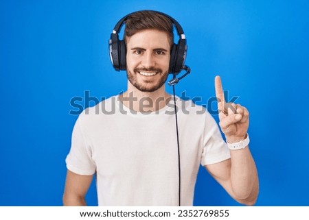 Hispanic man with beard listening to music wearing headphones showing and pointing up with finger number one while smiling confident and happy. 