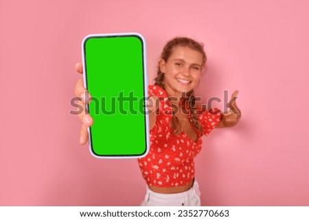 Young smiling optimistic Caucasian woman holds phone with green screen in hand and points to display of gadget with smile offering to place your promotional offer stands in pink studio.