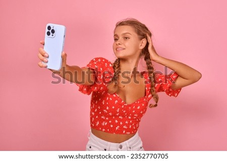 Young attractive blonde Caucasian woman poses for selfie and stretches out hand with mobile phone to take cool photo for model portfolio or dating site account stands on plain pink background.