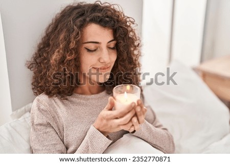 Young hispanic woman smelling aromatic candle sitting on bed at bedroom Royalty-Free Stock Photo #2352770871