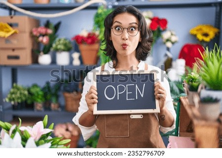 Young hispanic woman working at florist holding open sign making fish face with mouth and squinting eyes, crazy and comical. 