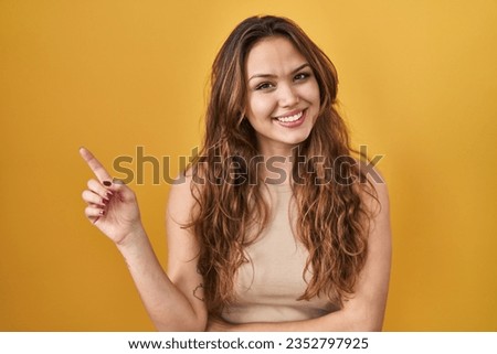 Young hispanic woman standing over yellow background with a big smile on face, pointing with hand and finger to the side looking at the camera. 