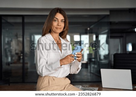 European businesswoman CEO holding smartphone using fintech application sitting on sofa in modern office. Smiling Latin Hispanic mature adult professional business woman using mobile phone cellphone.  Royalty-Free Stock Photo #2352826879