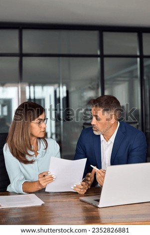 Mature 50s age Latin business man mentoring mid age European business woman discussing project with documents in office. Colleagues, group of partners, professional business people working together. Royalty-Free Stock Photo #2352826881