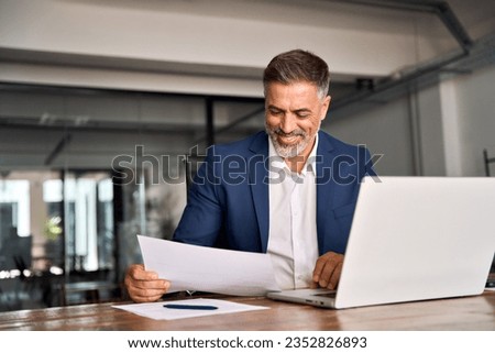 Smiling Latin or Indian male business man accountant analyst holding documents, work at laptop computer doing online trade market tech research. Focused Hispanic businessman with paperwork in office. Royalty-Free Stock Photo #2352826893