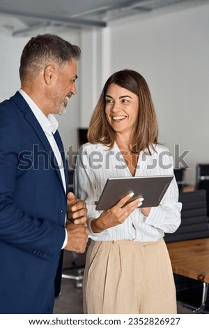 Vertical portrait of mature Latin businessman and European businesswoman discussing project on tablet in office. Two diverse partners colleagues of confident professional businesspeople work together. Royalty-Free Stock Photo #2352826927