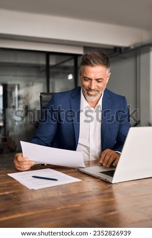 Smiling Latin or Indian male business man accountant analyst holding documents, work at laptop computer doing online trade market tech research. Hispanic businessman with paperwork in office, vertical Royalty-Free Stock Photo #2352826939