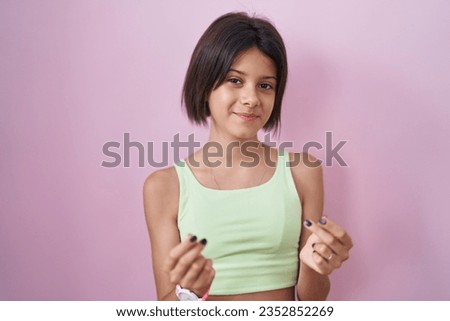 Young girl standing over pink background doing money gesture with hands, asking for salary payment, millionaire business 