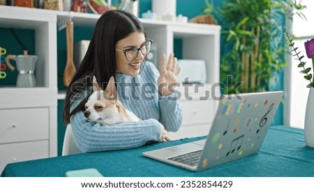 Young hispanic woman with chihuahua dog doing a video call waving to the laptop at dinning room