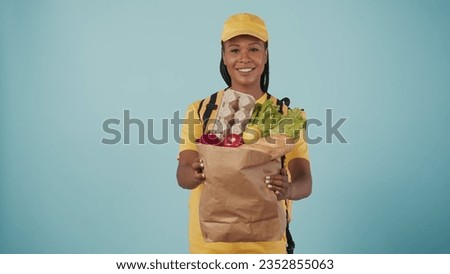 Delivery woman in uniform with portable refrigerator giving paper bag with grocery products. Isolated on blue background.