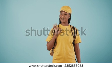 Delivery woman in yellow uniform with portable backpack refrigerator shows ok sign. Isolated on blue background.