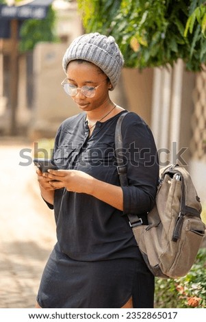 image of beautiful young african student enjoying social media outdoor- black lady with back pack holding smart phone