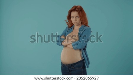 A redhaired pregnant woman gets upset, offended. A woman with her arms crossed on her chest stands in the studio on a blue background. Pregnancy and emotions, changeable mood, hormones.