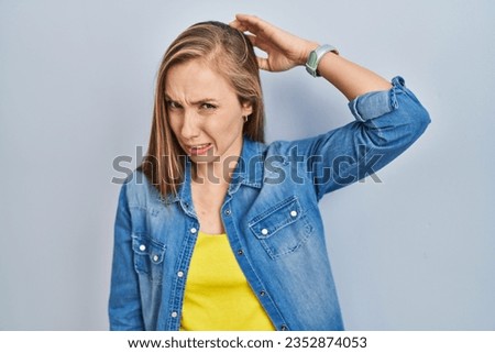 Young blonde woman standing over blue background confuse and wondering about question. uncertain with doubt, thinking with hand on head. pensive concept. 