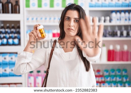 Young brunette woman shopping at pharmacy drugstore holding pills with open hand doing stop sign with serious and confident expression, defense gesture 