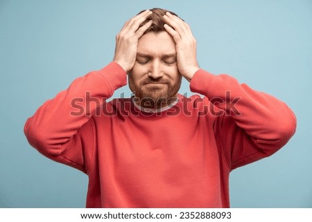 Irritated man with closed eyes holds head feeling discomfort due to headache on blue wall background. Concept of illness and suffering from unpleasant sensations. Sad bearded male regrets a mistake Royalty-Free Stock Photo #2352888093