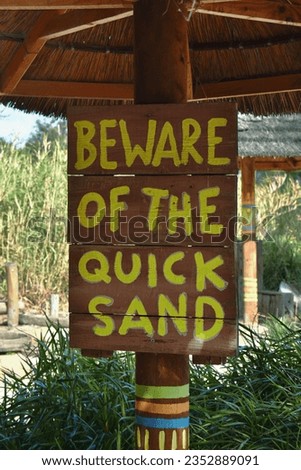 BEWARE OF THE QUICK SAND SIGNAGE - Handpainted yellow text words on an old rustic wooden planks quick sand signpost with a sandy and leafy green background