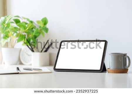 Digital tablet with blank display, coffee cup and potted plant on white table. Simple workplace.