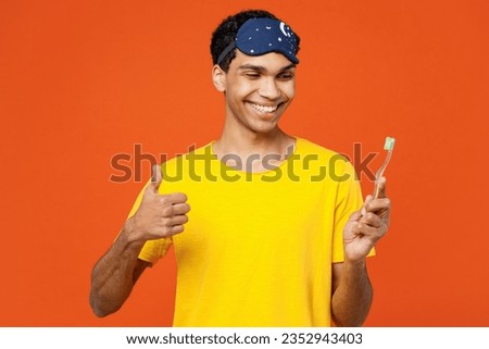 Calm smiling young man wear pyjamas jam sleep eye mask rest relax at home brushing teeth show thumb up isolated on plain orange background studio portrait. Good mood night nap, daily routine concept