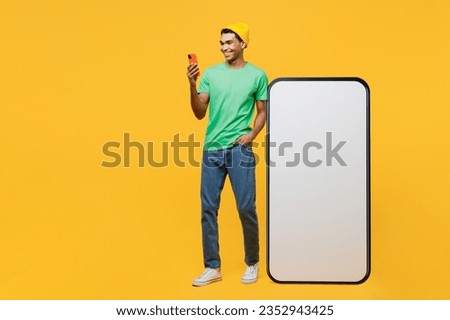 Full body smiling happy fun young man he wears casual clothes green t-shirt hat big huge blank screen mobile cell phone smartphone with area using mobile cell phone isolated on plain yellow background