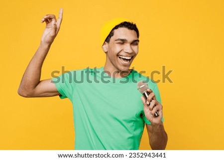 Young man of African American ethnicity he wears casual clothes green t-shirt hat sing song in microphone point index finger up isolated on plain yellow background studio portrait. Lifestyle concept