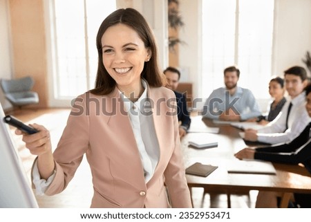Smiling young Caucasian businesswoman talk interact with diverse businesspeople at office meeting, happy female coach speaker make flip chart presentation for employees at briefing, training concept Royalty-Free Stock Photo #2352953471