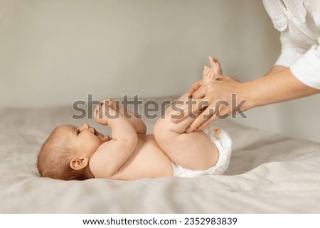 Constipation in babies. Mother doing gymnastics with her newborn child to relieve gases, moving kid's legs, infant lying on bed, side view, copy space