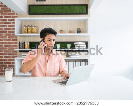 Asian traveller man confirm booking with airline call center by smartphone and check flight route and date by laptop together on kitchen home multitasking in the morning Royalty-Free Stock Photo #2352996333
