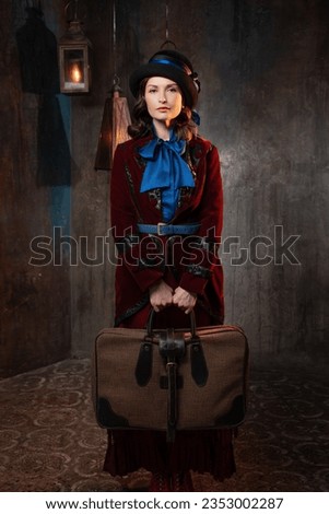 A stylish lady in a burgundy old-fashioned suit with a hat and a valise. Brunette in a retro style suit Royalty-Free Stock Photo #2353002287
