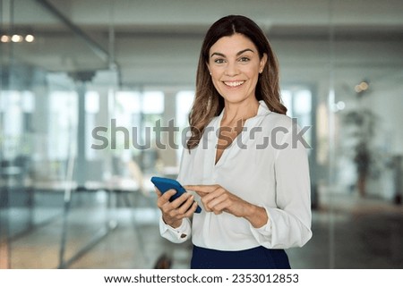 Happy mature professional business woman using cell phone at office, portrait. Smiling mid age 45 years old businesswoman executive standing at work lobby holding smartphone looking at camera.