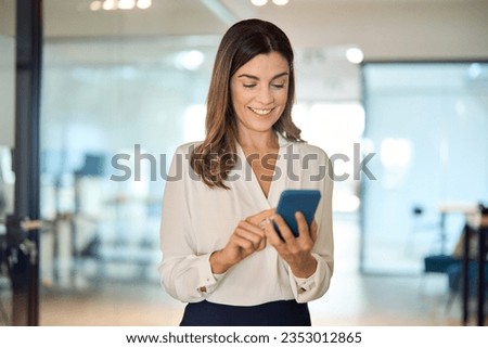 Smiling mid aged business woman executive using smartphone in office. Happy elegant mature female employee or professional bank sales manager looking at cell mobile phone tech standing in lobby.