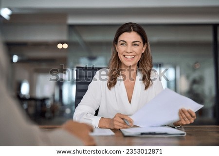 Smiling mature business woman hr holding cv document at job interview. Happy mid aged professional banking manager or lawyer consulting client sitting at workplace in corporate office meeting. Royalty-Free Stock Photo #2353012871
