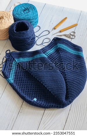 Crochet market bag with crochet needle and yarn balls on white wood background. Handmade and craft. Navy blue color crochet granny square bag.
