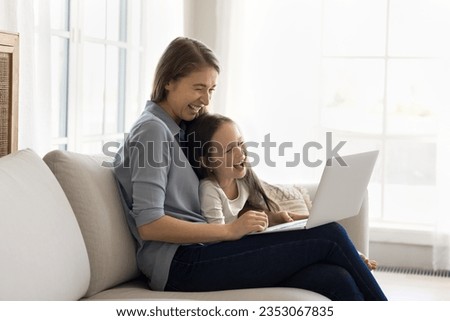 Happy joyful mom and cheerful kid watching funny movie, educational cartoon on laptop, smiling, laughing, resting on sofa at home, enjoying domestic wireless internet communication