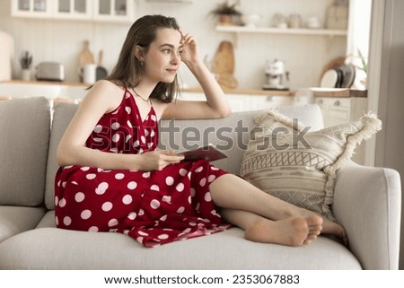 Positive dreamy young adult girl holding digital gadget, enjoying leisure, relaxing on home couch, looking at window away with thoughtful face, thinking, dreaming, smiling at good plans, thoughts