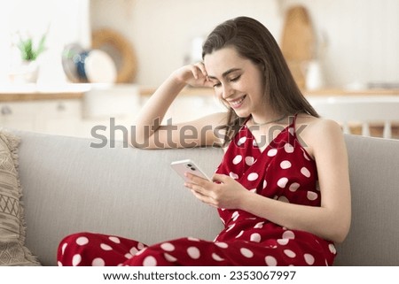 Happy beautiful young woman using smartphone for online shopping on Internet fashion stores, holding cellphone, resting on home sofa, smiling, laughing, enjoying Internet communication technology