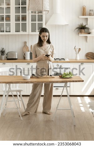 Happy beautiful young adult chef girl using mobile phone over kitchen table with organic food ingredients, reading recipe, ordering products, typing message, making salad, slicing vegetables