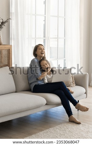 Excited cheerful mother cuddling pretty adorable kid on home sofa, using laptop, digital gadget for wireless online connection, watching movie, laughing, shouting, having fun with little girl
