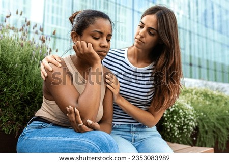 Young woman consoling and comforting upset and depressed African American female friend in park outdoors. Royalty-Free Stock Photo #2353083989