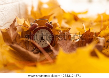 Alarm clock on table and beautiful autumn leaves on blurred background, space for text. Time change concept Royalty-Free Stock Photo #2353124371