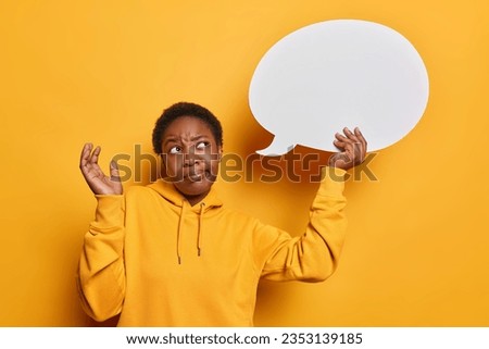 Photo of hesitant dark skinned young woman shrugs shoulders concentrated at white speech bubble for your promotion dressed in casual sweatshirt isolated over yellow background feels doubtful