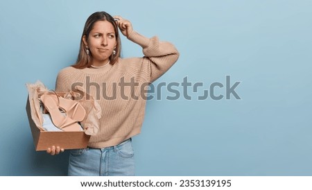 Studio shot of thoughtful woman scratches head looks pensively aside thinks which outfit to wear to fit shoes gets dressed for special occasion looks doubtful aside isolated over blue background