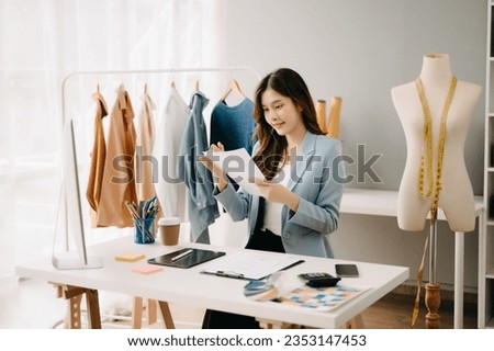 Successful fashion designer. Attractive young asian woman with smile while standing in workshop. Beautiful owner business woman working and holds tablet, laptop and smartphone in studio.
